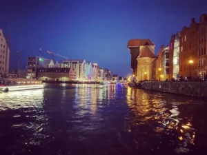 Nocna panorama nabrzeża w Gdańsku z podświetlonymi budynkami, zabytkowym Żurawiem oraz odbiciami świateł w wodzie. Luksusowe łodzie i statki dodają uroku wieczornej scenerii.