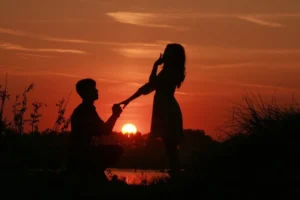 Romantyczna scena oświadczyn o zachodzie słońca – mężczyzna klęka na jedno kolano, trzymając rękę ukochanej, podczas gdy słońce zachodzi za ich sylwetkami, tworząc piękne cienie na tle nieba